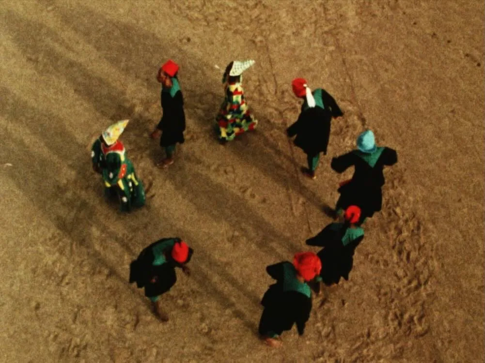 Overhead film still of figures in vivid head coverings crossing a sandy landscape in Hussein Shariffe’s work