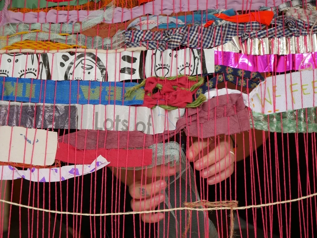 Hands weaving colourful fabric, foil and paper through the bright pink warp of a loom