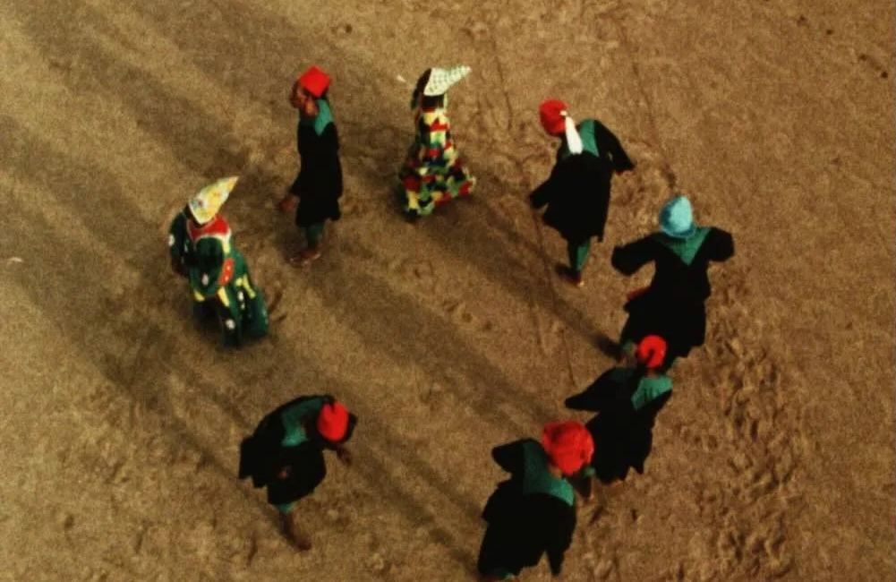 Overhead film still of figures in vivid head coverings crossing a sandy landscape in Hussein Shariffe’s work